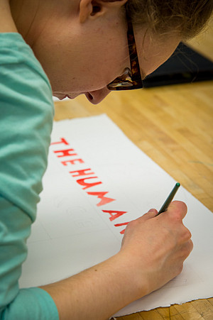 Student finishing a sign that says THE HUMANITIES in reddish-orange block letters