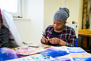 Student sitting at a table with paintings on it, using a pair of scissors in the Art Studio