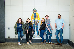 Students standing in the Second Avenue Subway Station in front of a wall that has a painting of a man on it